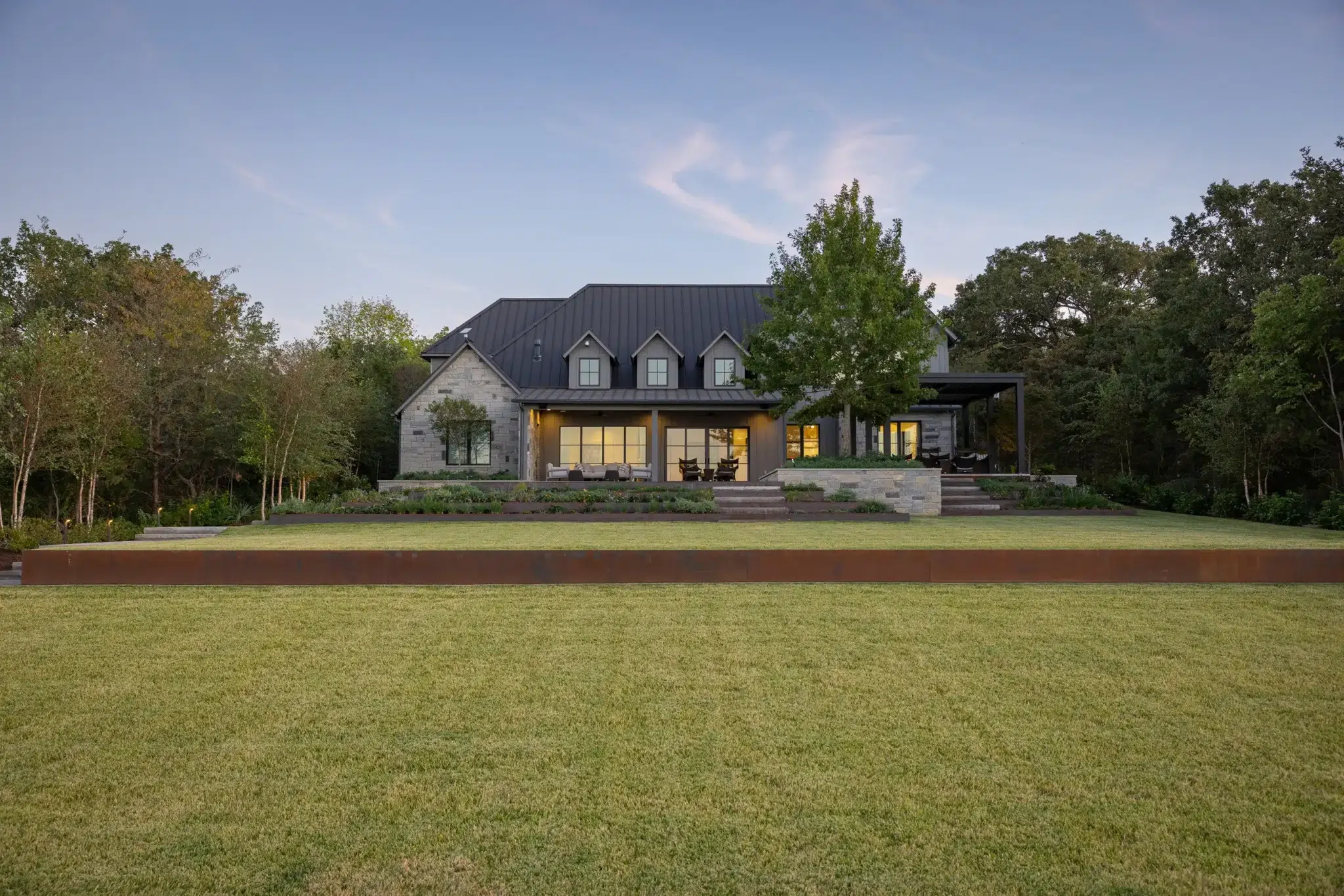 Rear elevation of Deer Valley home at Cedar Creek Lake at dusk with lit interior, steel retaining wall, and turf lawn designed by Landvisions